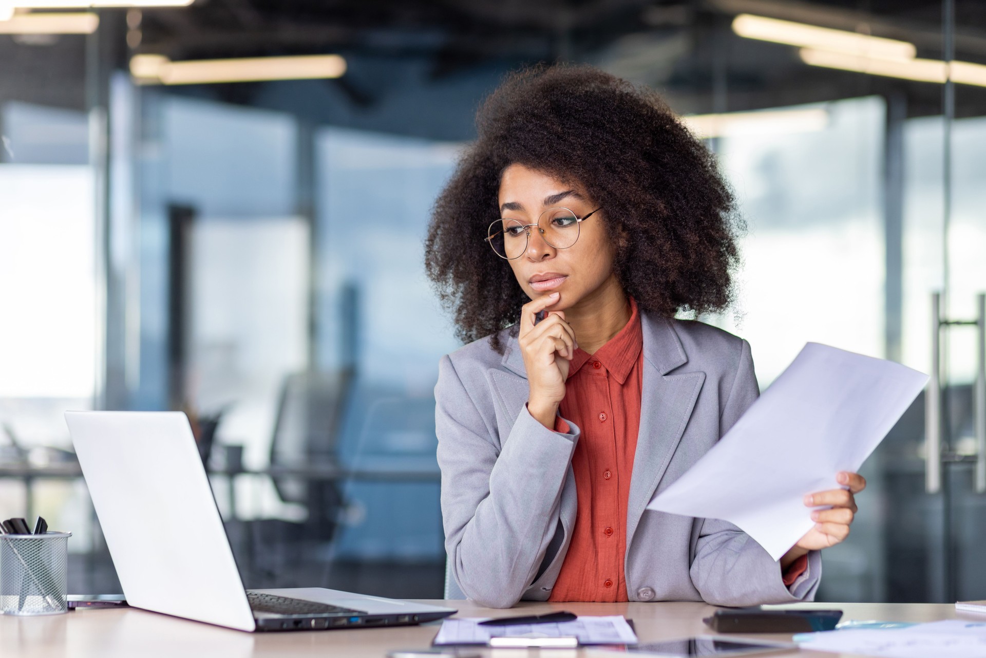 Focused project manager with curly hair holding document and looking with hesitation at computer screen. Suspicious woman checking accuracy of printed report and comparing with electronic version