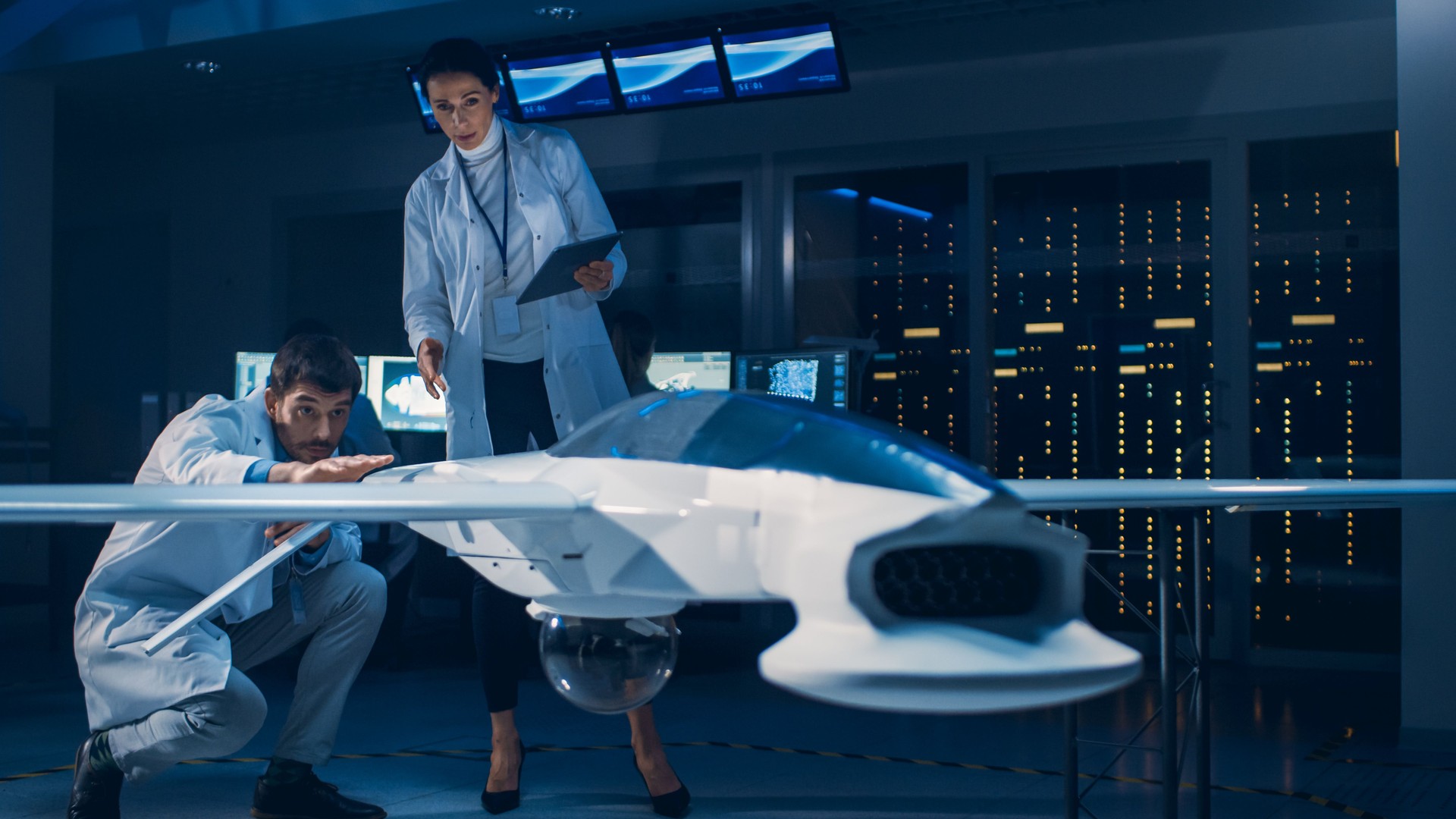 Meeting of Aerospace Engineers Working On Unmanned Aerial Vehicle / Drone Prototype. Aviation Scientists in White Coats Talking. Commercial Aerial Surveillance Aircraft in Industrial Laboratory